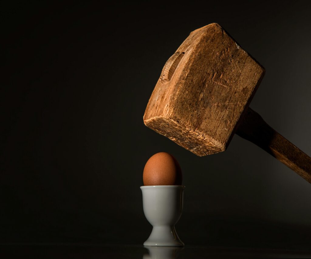 A wooden hammer poised above a fragile egg in a dramatic still life composition.