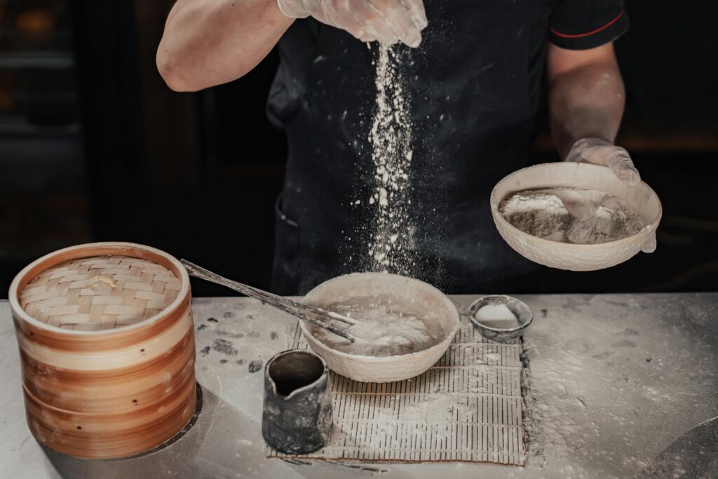 Chef sifting flour into a bowl, preparing dough in a rustic kitchen setting.